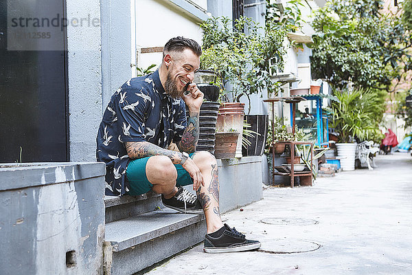 Junger Mann sitzt auf der Türschwelle in einer Wohngasse und telefoniert mit einem Smartphone  Shanghai Französische Konzession  Shanghai  China