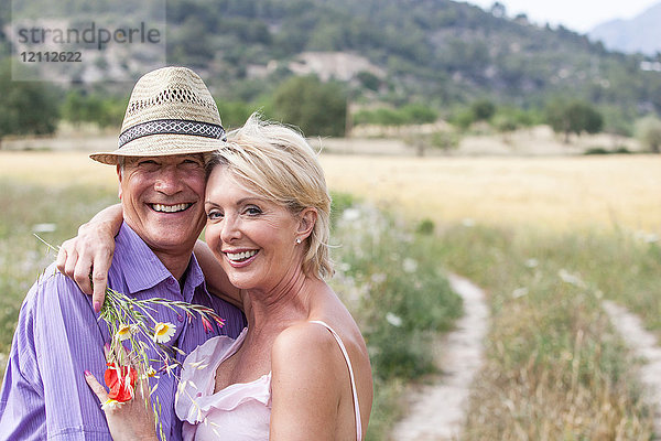 Paar im Feld mit Blumenstrauß  die sich umarmen und lächelnd in die Kamera blicken