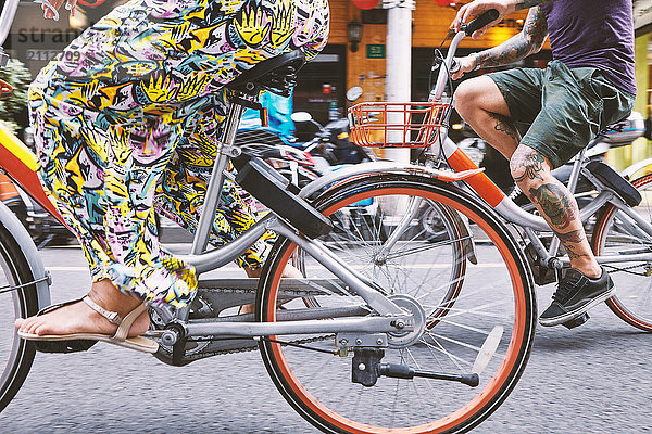 Taillenansicht eines multiethnischen Hipster-Paares  das mit dem Fahrrad die Straße entlang fährt  Schanghai Französische Konzession  Schanghai  China