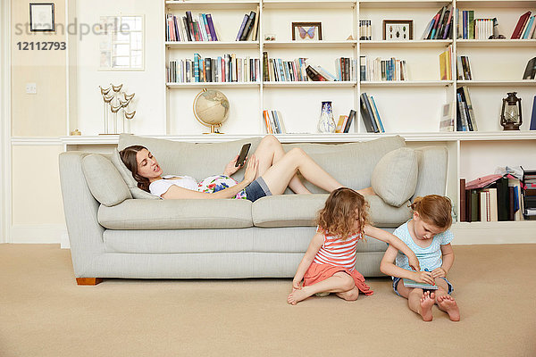 Schwangere Frau auf Sofa liegend mit Smartphone und Töchter auf Bodenzeichnung sitzend