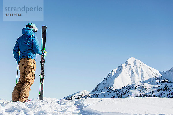 Skifahrer steht im Schnee  hält Skier in der Hand  schaut auf den Berg  Rückansicht