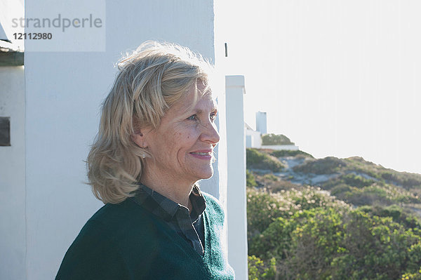 Glückliche blonde  ältere Frau  die von einem sonnenbeschienenen Haus an der Küste nach draußen schaut  Kapstadt  Westkap  Südafrika