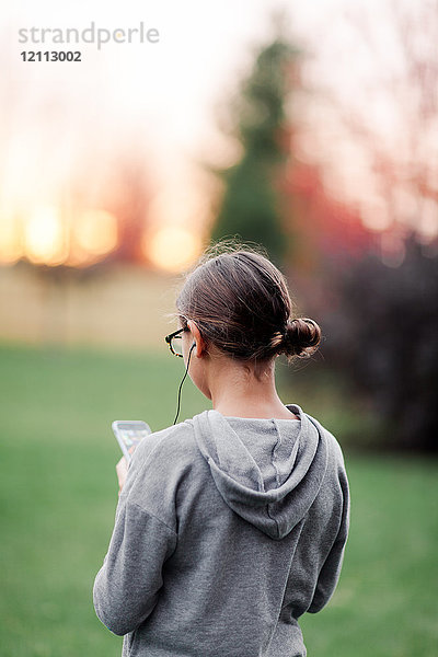 Rückansicht eines Mädchens  das über Kopfhörer Musik hört und auf ein Smartphone im Garten schaut