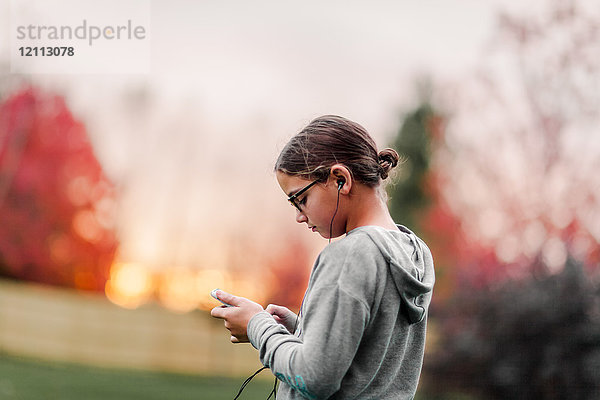 Seitenansicht eines Mädchens  das über Kopfhörer Musik hört und ein Smartphone im Garten betrachtet