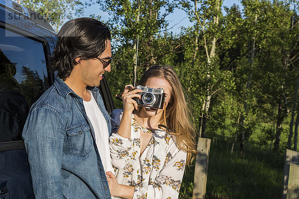 Junges Paar mit Kamera  das vor seinem Fahrzeug steht  während die junge Frau in der Ferne ein Foto macht; Edmonton  Alberta  Kanada