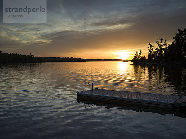 Sonnenuntergang über einem ruhigen See und silhouettierte Bäume entlang der Uferlinie mit einem Steg und einer Leiter im Vordergrund; Lake of the Woods  Ontario  Kanada