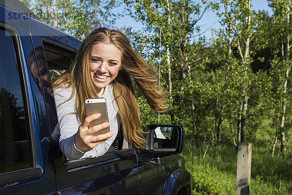 Eine schöne junge Frau mit langen blonden Haaren  die aus einem Fahrzeugfenster schaut und mit ihrem Smartphone ein Selbstporträt aufnimmt; Edmonton  Alberta  Kanada