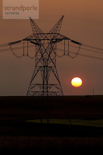 Silhouette eines großen Strommastes aus Metall mit Stromleitungen und orangefarbenem Sonnenball; Blackie  Alberta  Kanada