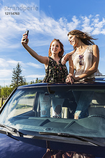 Zwei junge Frauen auf einem Roadtrip stehen auf dem Sonnendach eines Fahrzeugs und machen ein Selbstporträt mit einem Smartphone; Edmonton  Alberta  Kanada