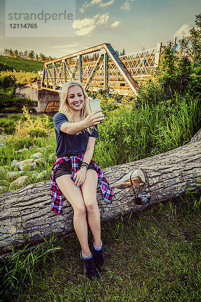 Eine junge Frau sitzt auf einem Baumstamm in einem Park und macht mit ihrem Handy ein Selbstporträt mit einer Brücke und einem Fluss im Hintergrund; Edmonton  Alberta  Kanada