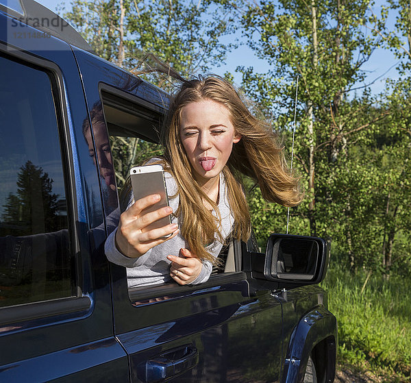 Eine schöne junge Frau mit langen blonden Haaren  die aus dem Fenster eines Fahrzeugs schaut und ein albernes Selbstporträt mit ihrem Smartphone aufnimmt  während sie der Kamera die Zunge herausstreckt; Edmonton  Alberta  Kanada