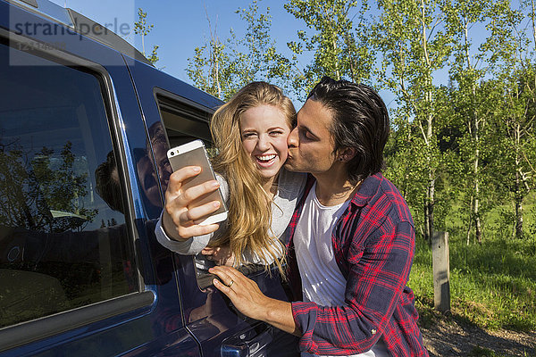 Ein junges Paar macht ein Selbstporträt mit seinem Smartphone an seinem Fahrzeug  während er sie auf die Wange küsst; Edmonton  Alberta  Kanada