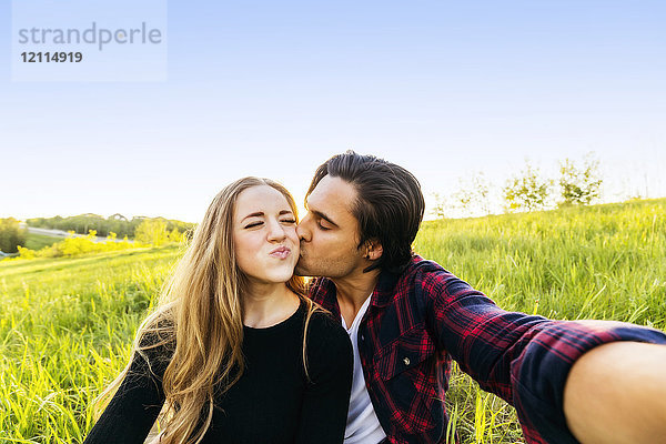 Ein junges Paar sitzt auf einem Feld und nimmt ein Selbstporträt auf  während der junge Mann die junge Frau auf die Wange küsst; Edmonton  Alberta  Kanada