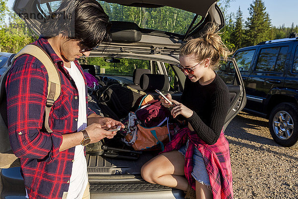 Junges Paar  das auf dem Rücksitz seines vollgepackten Fahrzeugs steht  die hintere Tür geöffnet hat und beide ihre Smartphones benutzen; Edmonton  Alberta  Kanada