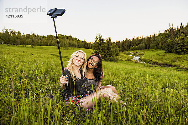 Zwei Freundinnen sitzen auf einer Wiese und posieren für ein Selbstporträt mit einem Selfie-Stick und einem Smartphone; Edmonton  Alberta  Kanada