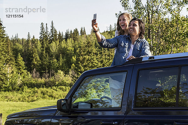 Junges Paar  das Selbstporträts mit einem Mobiltelefon macht  während es auf dem Schiebedach seines Fahrzeugs steht; Edmonton  Alberta  Kanada