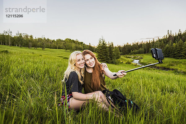 Zwei Freundinnen sitzen auf einer Wiese und posieren für ein Selbstporträt mit einem Selfie-Stick und einem Smartphone bei Sonnenuntergang; Edmonton  Alberta  Kanada