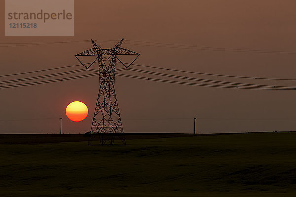 Silhouette eines großen Strommastes aus Metall mit Stromleitungen und orangefarbenem Sonnenball; Blackie  Alberta  Kanada