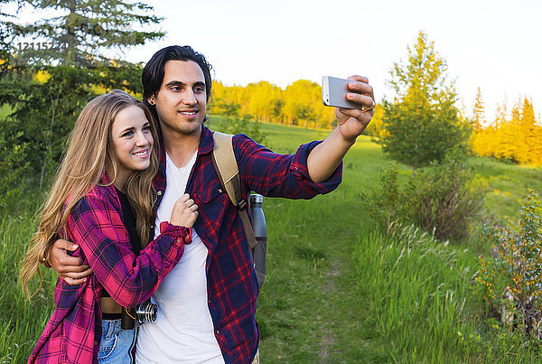 Junges Paar in einem Park  das bei Sonnenuntergang mit seinem Mobiltelefon für ein Selbstporträt posiert; Edmonton  Alberta  Kanada