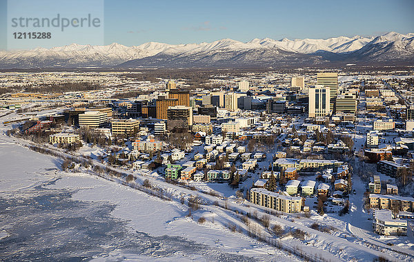 Luftaufnahme von Schnee  der das Meereis an den gefrorenen Ufern der Innenstadt von Anchorage bedeckt  die Chugach Mountains in der Ferne hinter den Bürogebäuden und Hotels  Cook Inlet im Vordergrund  Süd-Zentral-Alaska im Winter; Anchorage  Alaska  Vereinigte Staaten von Amerika