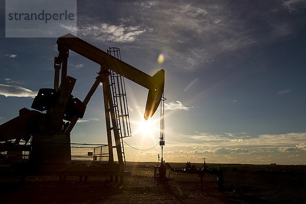 Silhouette eines Pumpjacks im Gegenlicht der untergehenden Sonne in den Prärien von Alberta mit blauem Himmel und Wolken  westlich von Airdrie; Alberta  Kanada