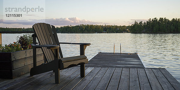 Ein hölzerner Adirondack-Stuhl steht auf einem Steg an einem ruhigen See bei Sonnenuntergang; Lake of the Woods  Ontario  Kanada