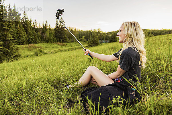 Eine junge Frau mit langen blonden Haaren macht ein Selbstporträt mit einem Selfie-Stick und einem Smartphone  während sie auf einer Wiese in einem Park sitzt; Edmonton  Alberta  Kanada
