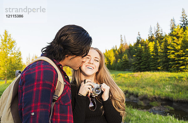 Junges Paar geht zusammen in einem Stadtpark spazieren  die junge Frau hält eine Kamera und der junge Mann küsst sie auf die Wange; Edmonton  Alberta  Kanada