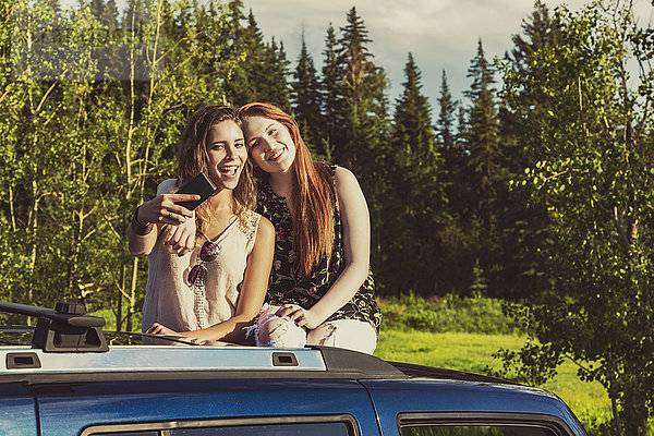 Zwei junge Frauen auf einem Roadtrip stehen auf dem Sonnendach eines Fahrzeugs und machen ein Selbstporträt mit einem Smartphone; Edmonton  Alberta  Kanada