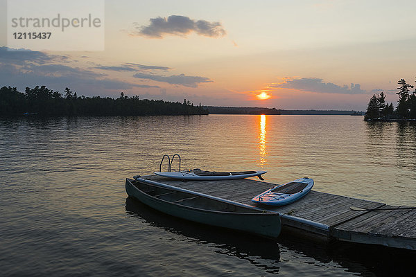 Sonnenuntergang über einem ruhigen See mit einem Steg  Kanu und Paddelbrettern im Vordergrund; Lake of the Woods  Ontario  Kanada