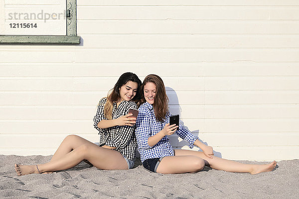 Zwei Mädchen im Teenageralter sitzen im Sand vor einem Gebäude und schauen gemeinsam auf ihre Smartphones  Woodbine Beach; Toronto  Ontario  Kanada
