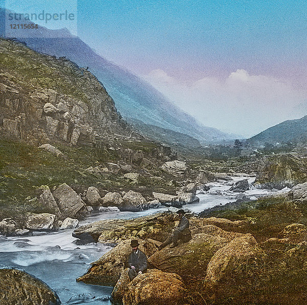 Diapositiv mit Laterna Magica um 1900  handkoloriert  erstellt 1887. Eine Tour durch Nordwales. 28 Llanberis Pass - Seen in der Ferne - Diese Ansicht bietet sich auf halber Höhe des Passes  wenn man nach rechts von der Straße abbiegt  an der Seite eines tosenden Wildbachs  der zum Llyn Peris hinunterstürzt. Hier befinden wir uns inmitten einer grandiosen Landschaft. Auf allen Seiten liegen große Felsmassen  die sich in einem vergangenen Zeitalter von den darüber liegenden Bergen gelöst haben. Diese monströsen Fragmente  die an den steilen Hängen der Berge verstreut sind  verstärken die Unheimlichkeit der Szene  die jedoch durch den Blick auf die Seen  die ruhig und friedlich am Ende des düsteren Tals liegen  noch verstärkt wird.