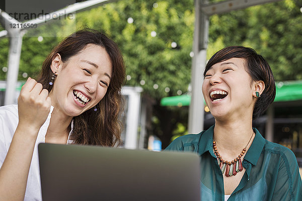 Zwei Frauen mit schwarzen Haaren in grün-weißem Hemd sitzen lachend vor dem Laptop am Tisch in einem Straßencafé.