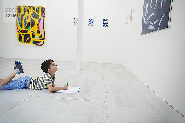 Junge mit kurzen schwarzen Haaren  der in der Kunstgalerie mit Stift und Papier auf dem Boden liegt und moderne Malerei betrachtet.