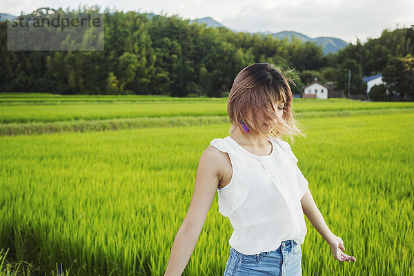 Eine junge Frau in weißem Hemd und Jeans mit ausgestreckten Händen steht auf freiem Feld an Reisfeldern.