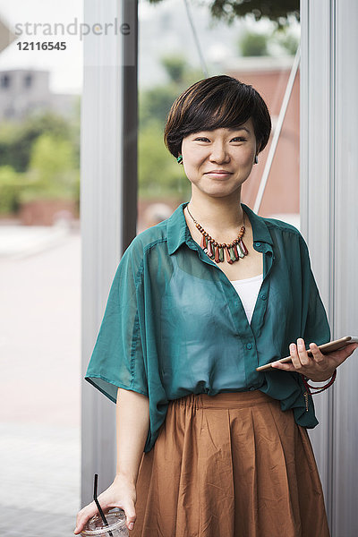 Frau mit schwarzen Haaren und grünem Hemd steht im Freien  hält ein digitales Tablett in der Hand und lächelt in die Kamera.