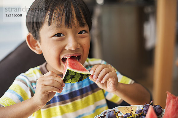 Nahaufnahme eines Jungen mit schwarzen Haaren  der ein gestreiftes T-Shirt trägt und eine Scheibe Wassermelone isst.