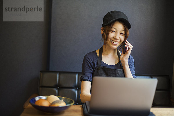 Frau arbeitet in einer Bäckerei  trägt eine Baseballmütze  sitzt am Tisch vor einem Laptop und benutzt ein Mobiltelefon.