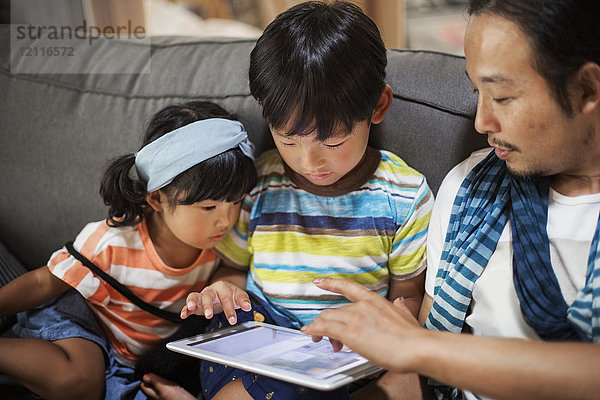 Mann  Junge und junges Mädchen sitzen auf einem grauen Sofa und schauen auf ein digitales Tablett.