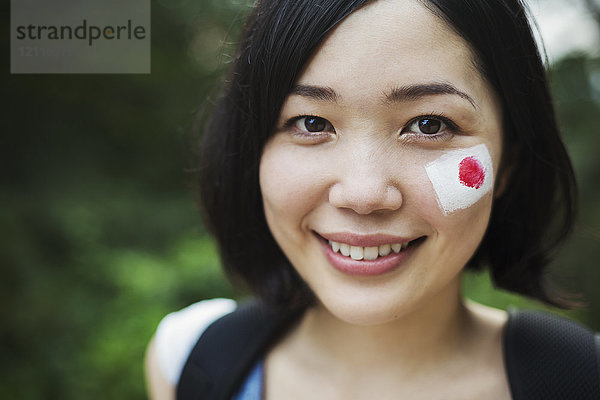 Porträt einer jungen Frau mit schwarzen Haaren  japanische Flagge auf die Wange gemalt  lächelt in die Kamera.