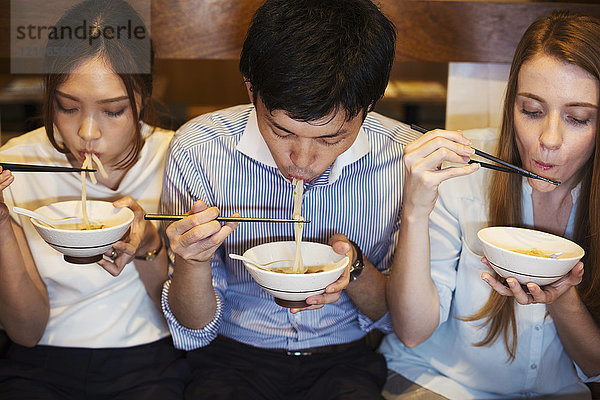 Hochwinkelaufnahme von drei Personen  die nebeneinander an einem Tisch in einem Restaurant sitzen und mit Stäbchen aus Schüsseln essen.