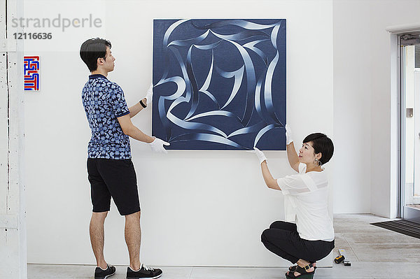 Mann in blauem Hemd und Frau mit weißem Hemd hängen modernes Gemälde an weißer Wand in Kunstgalerie.