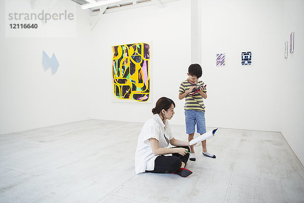 Frau mit schwarzen Haaren in weißem Hemd mit Stift und Papier auf dem Boden in der Kunstgalerie sitzend  neben ihr steht ein Junge.