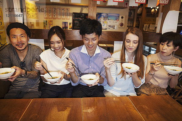 Fünf Personen sitzen nebeneinander an einem Tisch in einem Restaurant und essen mit Stäbchen aus Schalen.