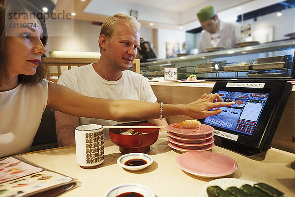 Junger Mann und Frau sitzen an einem Tisch in einem asiatischen Fast-Food-Restaurant  essen Sushi und schauen auf einen Touchscreen auf ihrem Tisch.