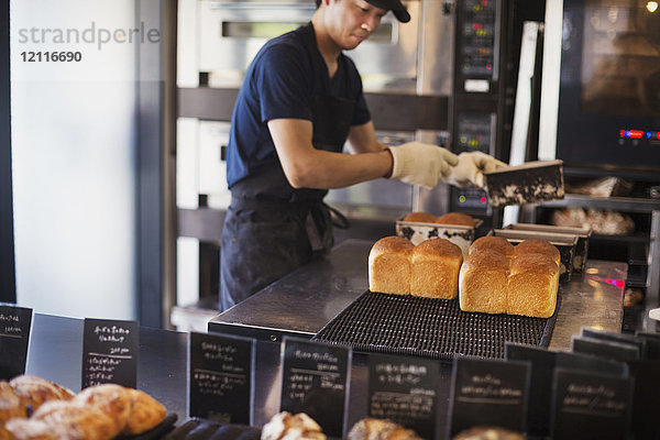 Mann  der in einer Bäckerei arbeitet und frisch gebackene Brotlaibe auf ein Tablett legt.