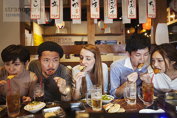 Fünf Personen sitzen nebeneinander an einem Tisch in einem Restaurant und essen von Spießen.