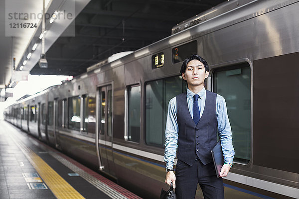 Geschäftsmann in blauem Hemd und Weste steht auf dem Bahnsteig des Bahnhofs und hält ein digitales Tablett und eine Aktentasche.