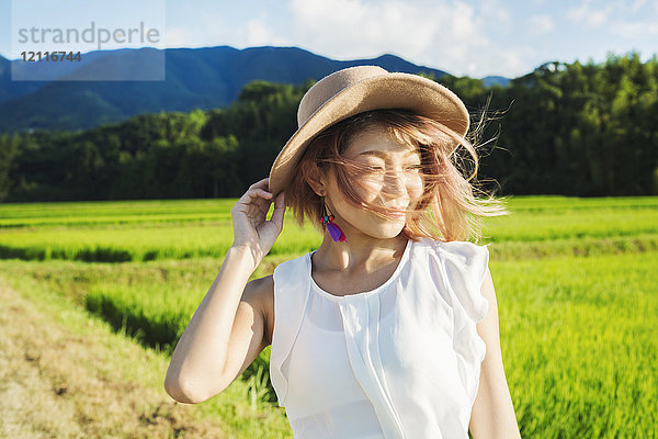 Eine junge Frau hält ihren Hut im Wind  vorbei an Reisfeldern mit grünen Trieben und Berglandschaft.