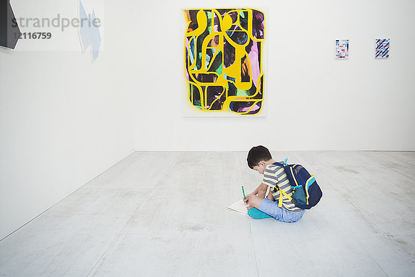 Junge mit kurzen schwarzen Haaren und Rucksack  der mit Stift und Papier auf dem Boden in der Kunstgalerie sitzt und moderne Malerei betrachtet.